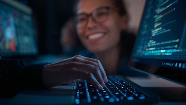 Smiling Female Developer Typing on Backlit Keyboard with Code on Monitor in Blue Glow
