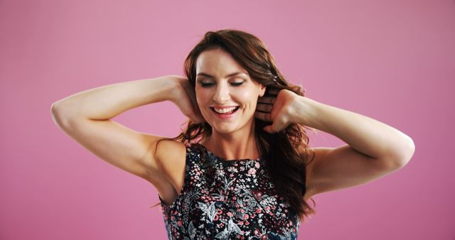 This image depicts a cheerful woman in a floral dress stretching with a bright smile on her face against a pink background. It exudes positive energy and relaxation, making it suitable for use in advertisements for beauty, fashion, lifestyle blogs, and wellness content. The bright pink background adds a vibrant touch, making it perfect for promotional materials targeting a young and trendy audience.