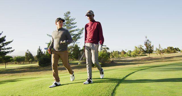 Two male golfers are walking together on a well-manicured golf course on a sunny day. Both are holding golf clubs, wearing golf attire, and engaging in conversation. This image can be used for themes related to sports, leisure activities, men's friendship, outdoor lifestyles, and golfing events or promotions.