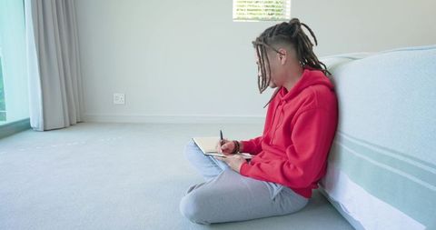 Mid-adult man journaling on bedroom floor wearing red hoodie and dreadlocks