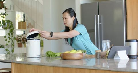 Asian woman composting vegetable scraps on modern kitchen island with tablet and produce