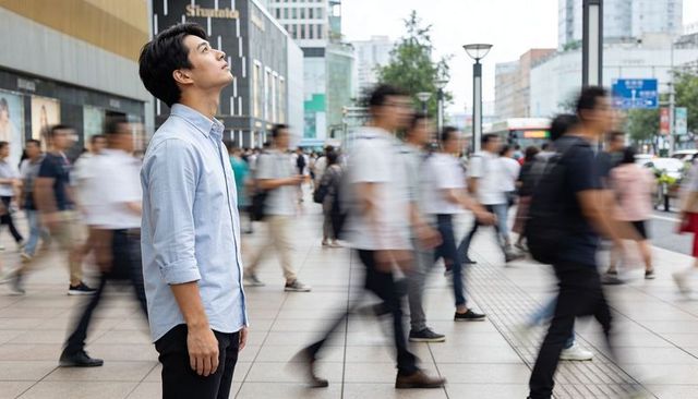 Young asian man standing in busy urban plaza while crowd rushing by, contemplative commuter