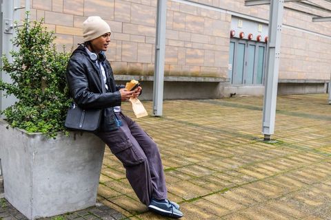 Young man sitting on planter with beanie and headphones holding sandwich in urban courtyard