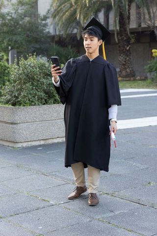 Asian graduate checking phone and holding diploma on university campus in cap and gown