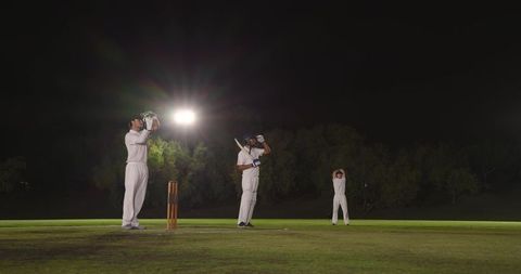 Night cricket under floodlights featuring batsman adjusting helmet and diverse fielders in white