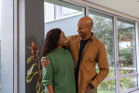 Diverse couple embracing by glass doors in modern apartment, cozy green sweater, natural light