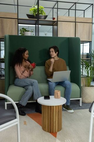 Diverse coworkers chatting on green booth, using laptop and holding mugs in modern office