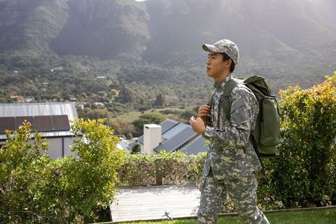 Asian soldier walking on suburban hillside carrying olive backpack overlooking valley