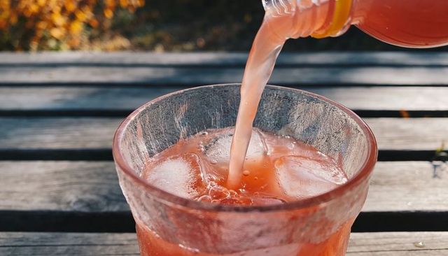 Pouring pink citrus drink into clear glass with ice on sunlit weathered wooden table