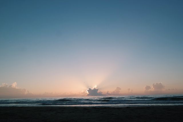 Serene beach sunrise with sun rays shining through clouds over calm ocean and wide horizon. Ideal for relaxation themes, inspirational quotes, travel blogs, and meditation content.