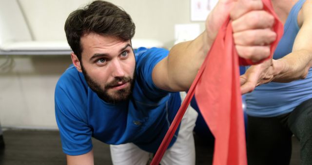 Man working out with red resistance band in a gym, demonstrating exercise technique and strength training. Useful for illustrating fitness tutorials, personal training, healthy lifestyle, and physical education content.