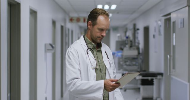 The image shows a male doctor in a hospital corridor, reviewing a medical chart. He is wearing a white coat with a stethoscope around his neck, indicating his medical profession. This scene can be used in promotional materials for medical and healthcare services, educational content on medical professions, or presentations emphasizing patient care and professional healthcare contexts.