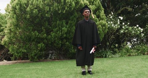 Graduate standing in cap and gown holding diploma on green lawn marking academic success