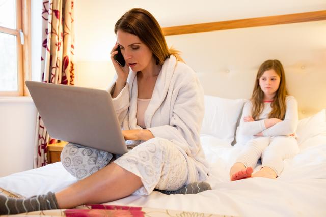 Mother using laptop with her upset daughter sitting on bed in bedroom