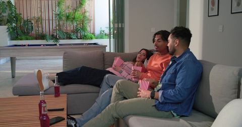 Three Friends Lounging on Grey Sectional Holding Popcorn and Bottles for Movie Night