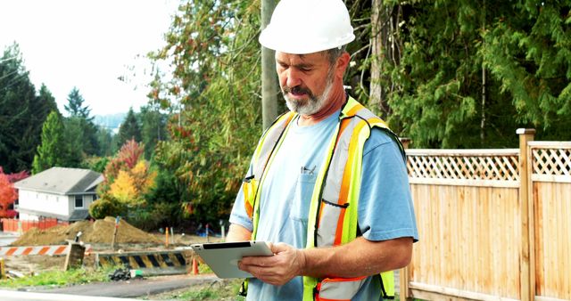Mature construction worker using a digital tablet at an outdoor job site, wearing a safety vest and hard hat. Ideal for use in articles or advertisements related to construction technology, site management, field engineering, worker safety, and project planning.