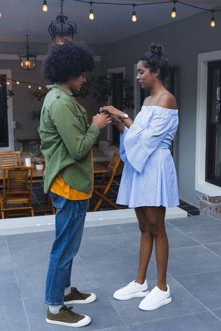 Young african american man placing ring on partner on romantic patio with string lights