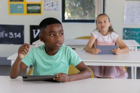 Black boy raising hand while holding tablet with classmate using tablet in modern classroom
