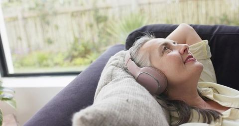 Senior woman reclining on sofa listening to music with pink wireless headphones by window