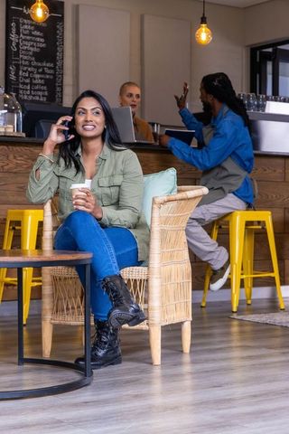 Indian woman sitting in wicker chair holding coffee and talking on phone in cozy cafe