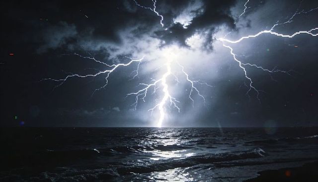 Lightning bolt branching across night sky, striking ocean and reflecting on stormy shoreline