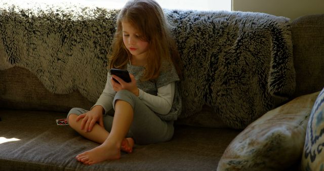 Young girl comfortably relaxing on a couch while using a smartphone. Her casual attire and serene environment indicate a typical scene of childhood leisure in a home setting. This can be used to depict themes of childhood, technology use among kids, home relaxation, family life, or modern lifestyle.
