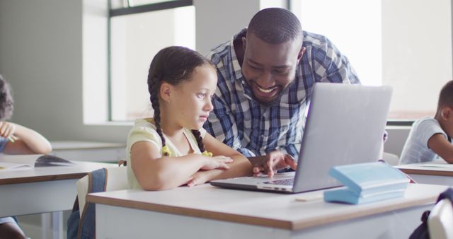 Image of happy african american male teacher helping caucasian girl with laptop. primary school education and teaching profession.