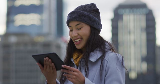 Young woman using tablet in winter cityscape, wearing cozy coat and beanie. Suitable for themes related to technology, urban lifestyle, winter fashion, digital communication.