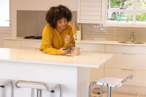 Young woman leaning on kitchen island checking smartphone while holding coffee mug