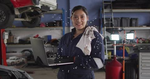 Female auto technician holding laptop while inspecting vehicle on lift in garage workshop