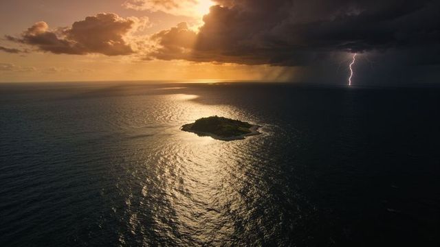 Isolated tropical islet glowing under stormy sunset with lightning striking over golden ocean reflec