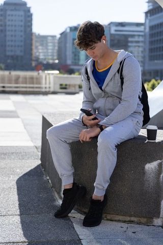 Young man checking smartphone on urban ledge wearing grey tracksuit and earbuds