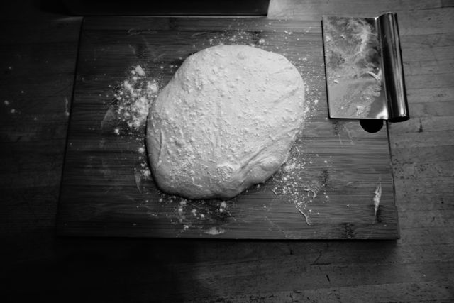 Rustic pizza dough is resting on a wooden board, lightly dusted with flour. Ideal for illustrating homemade baking, cooking tutorials, or kitchen scenes.