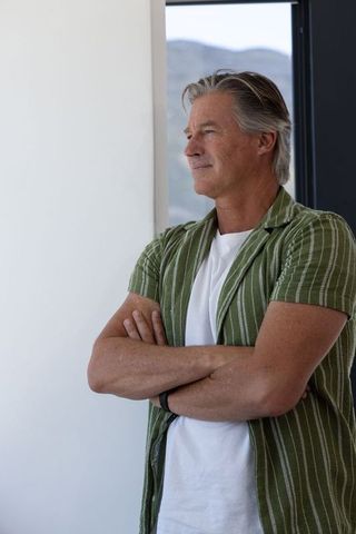 Mature man standing by window gazing out in modern sunlit home wearing green striped shirt