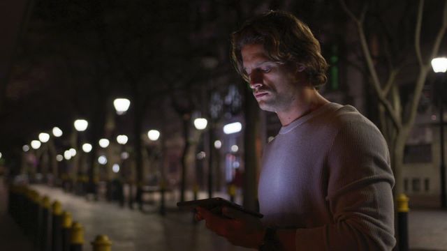 Young Caucasian man standing on an urban street at night, illuminated by street lights using a tablet and smiling. Ideal for technology, urban lifestyle, or nighttime city-living themes.