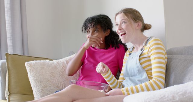 Two young girls are sitting on a couch and laughing while watching television. One girl is wearing a pink shirt and the other has on a striped yellow shirt with overalls. They have a glass bowl of snacks on their lap. This image can be used for themes related to friendship, leisure activities, and enjoying quality time together. Ideal for blogs, advertising, and social media posts promoting relaxation, bonding moments, and fun at home.