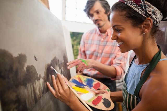Man assisting woman in painting during art class. Woman holding palette and brush, smiling while painting on canvas. Man providing guidance and support. Ideal for use in educational materials, art class promotions, creative workshops, and hobbyist communities.