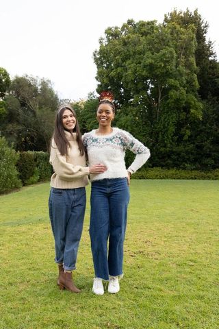 Two diverse women standing on garden lawn wearing sweaters and jeans smiling together