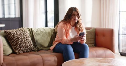 Young woman sitting on leather sofa scrolling smartphone in bright cozy living room