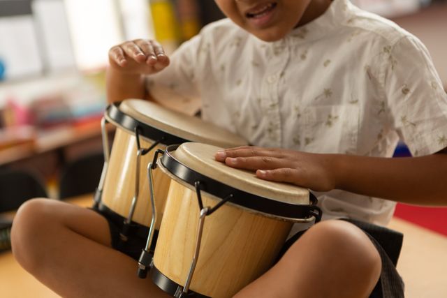 Biracial boy playing bongo drums, focused on music. He has short curly hair, wearing patterned shirt, unaltered