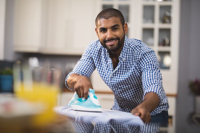 Young man in plaid shirt ironing clothes in a modern kitchen. Ideal for themes related to domestic chores, household tasks, modern lifestyle, and multitasking. Useful for advertisements, blogs, and articles about home life, gender roles, and daily routines.