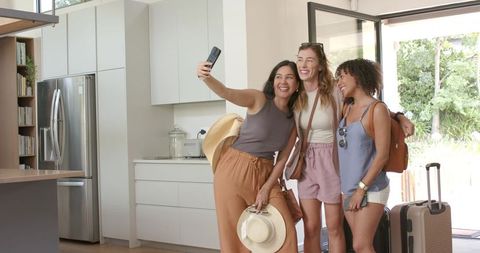 Multicultural friends posing for travel selfie in sunlit modern kitchen with luggage