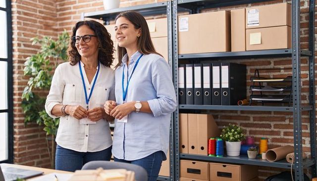 Two female coworkers smiling and planning in small business packaging studio with shelving