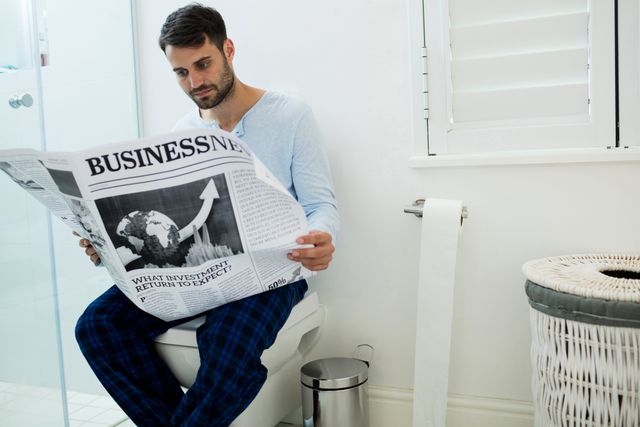 Man sitting on toilet seat reading a newspaper at home