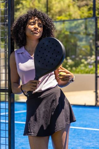 Confident sunlit padel player leaning on fence with black racket and yellow ball