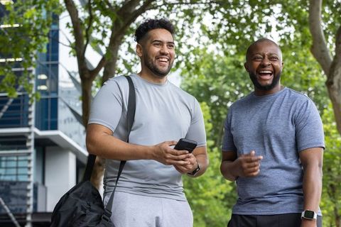 Two african american men laughing after workout in urban park with smartphone and gym bag
