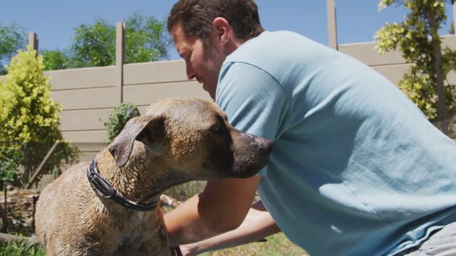 A Caucasian male volunteer wearing a blue shirt is caring for a dog in a paddling pool at an animal shelter under bright sunshine. This scene captures compassion and outdoor activity, making it perfect for use in content related to animal rescue, volunteering, outdoor pet care activities, or promotion of responsible pet ownership.