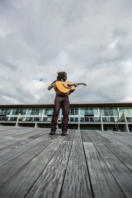 Man enjoying his time playing an acoustic guitar while standing on a wooden boardwalk against a backdrop of a modern glass building. The overcast sky adds a dramatic effect to the scene. Perfect for use in advertisements, music promotions, entertainment content, lifestyle articles, and modern urban life depictions.