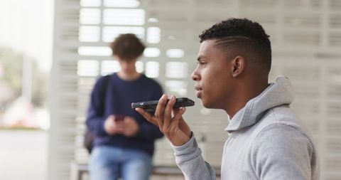 Young Man Speaking into Smartphone at Bus Shelter While Friend Browsing Phone, Commuter