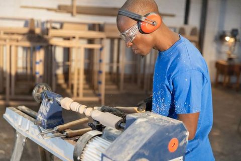 Young male woodworker shaping spindle on lathe wearing ear protection and goggles